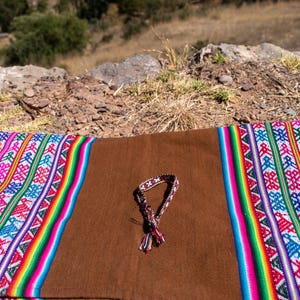Peut inclure: Une textile tissé coloré avec un centre marron et une bordure à motifs roses, bleus, verts et oranges. Un petit bracelet tissé de manière complexe avec des accents rouges et blancs repose sur le textile.