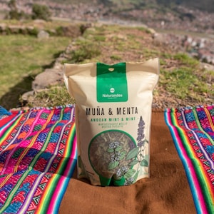 May include: A beige bag of "Muña & Menta" Andean mint and mint tea, with a green top, sits on a colorful woven textile. The bag features a circular image of the tea leaves and a botanical illustration. The background shows a hillside and a distant town.