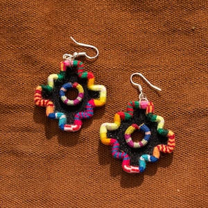 Puede incluir: Un par de pendientes colgantes con una base negra en forma de estrella de cuatro puntas y un recorte circular central. Los bordes y el círculo interior están bordados con hilo multicolor vibrante en patrones ondulados de rojo, amarillo, azul y verde.