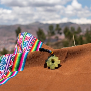 Puede incluir: Un colgante de piedra verde con un diseño en forma de cruz descansa sobre un textil marrón. Un textil tejido colorido con patrones geométricos y rayas arcoíris está drapeado sobre la tela marrón. El fondo presenta una cordillera bajo un cielo nublado.