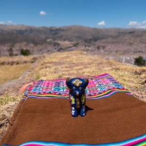 May include: A blue ceramic bull figurine with gold accents stands on a brown blanket with colorful woven patterns. The blanket is placed on a rocky surface, with a mountain range and blue sky in the background.