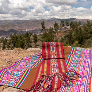 May include: A colorful textile display featuring traditional woven patterns. The textiles are primarily red, brown, and pink, with intricate designs in various colors. The scene is set outdoors with a mountain backdrop.