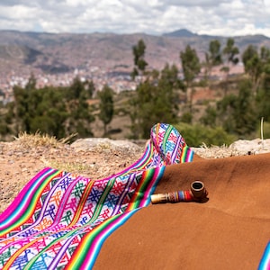 May include: A brown blanket with colourful woven trim and a small wooden pipe rests on a rocky surface. The blanket's trim features vibrant stripes and geometric patterns. The background shows a distant city and mountains under a cloudy sky.