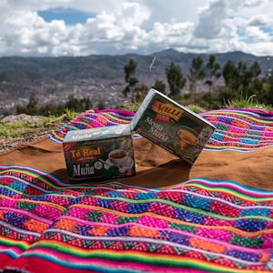 May include: Two boxes of "Té Real Muña" tea sit on a colorful woven blanket with a brown leather border. The boxes are green and white, with images of a cup of tea. The background features a mountain range and a cloudy sky.