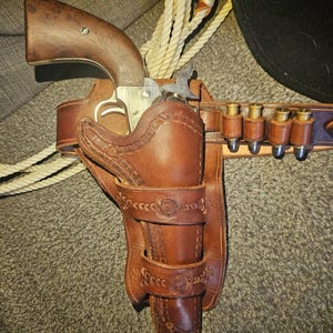 Könnte beinhalten: Ein braunes Lederholster im Vintage-Stil mit passendem Gürtel und Patronen. Das Holster ist für einen Revolver mit Holzgriff konzipiert. Der Gürtel hat Schlaufen für Patronen. Ein Seil und ein schwarzer Hut sind im Hintergrund zu sehen.