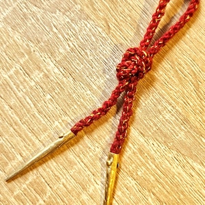 May include: A red bolo tie with gold accents. The braided cord is a deep red with gold metallic threads woven throughout. The tips are gold-colored metal, pointed and tapered. The tie is knotted at the center.