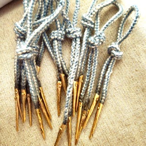 May include: A collection of blue and white braided cords with gold-colored metal tips. The cords are knotted at the top, creating loops. The tips are pointed and appear to be made of brass or a similar metal. The background is a textured, neutral-colored fabric.