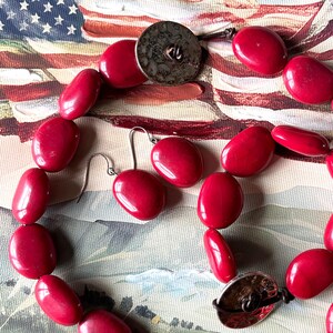 May include: A red beaded necklace and matching earrings set. The necklace features oval-shaped beads and a silver-tone clasp. The earrings are a simple oval shape. The jewelry is displayed against a backdrop of an American flag.