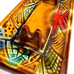 May include: A rectangular ceramic ashtray with a raised center section. The ashtray features a brown, orange, and yellow color scheme with a textured, patterned design. Accents of red, green, and blue are also present.