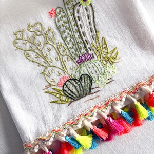 May include: White hand towel with an embroidered cactus design in shades of green, pink, and purple. The bottom edge features a colorful trim with neon pink, yellow, and blue tassels, adding a vibrant touch to the desert-themed textile.