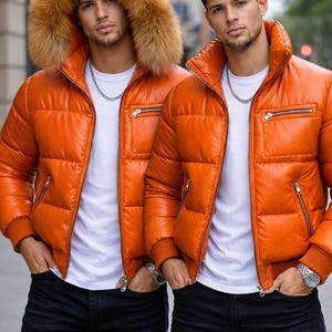 Puede incluir: Dos hombres con chaquetas acolchadas naranjas, vaqueros negros y camisetas blancas. Una chaqueta tiene una capucha forrada de piel. Ambos llevan collares de cadena plateada y relojes. Las chaquetas tienen bolsillos con cremallera.