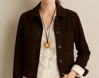 May include: A dark brown suede jacket with a collared neckline, button closure, and two front pockets. The jacket is worn over a white lace blouse and paired with white pants. A wooden pendant necklace is also visible.
