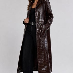 Peut inclure: Long manteau en similicuir marron avec une texture peau de crocodile. Le manteau a un col cranté, des poches et une ceinture. La personne porte un haut noir, un jean noir et des talons noirs.