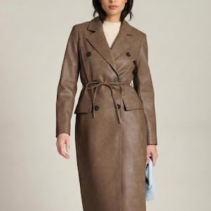 Può includere: Un trench coat marrone in ecopelle con un design doppiopetto, una cintura in vita e una gonna al ginocchio. Il cappotto presenta due tasche anteriori e un revers a lancia. La modella indossa un maglione color crema sotto.