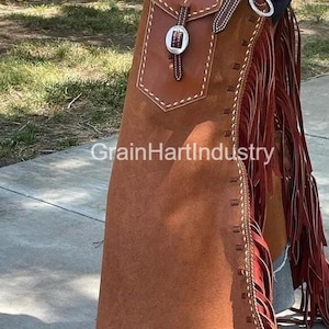May include: Brown leather chaps with a pocket, silver buckle, and decorative stitching. The chaps have long red fringe along the sides and the text "GrainHartIndustry".