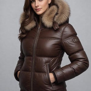May include: A brown leather puffer jacket with a fur-trimmed hood. The jacket has a zip-up front, pockets, and a ribbed hem. The model is wearing blue jeans. The jacket is designed for cold weather.
