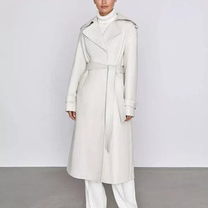 Puede incluir: Un abrigo largo tipo gabardina de color blanco roto con cinturón a juego, usado sobre un cuello alto y pantalones blancos. El abrigo presenta un diseño clásico con solapas y mangas largas. El conjunto es monocromático.