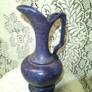 May include: A vintage, speckled, blue ceramic pitcher with a rounded body, a curved handle, and a spout. The pitcher has a bulbous base and a textured surface. The background is a white lace tablecloth.