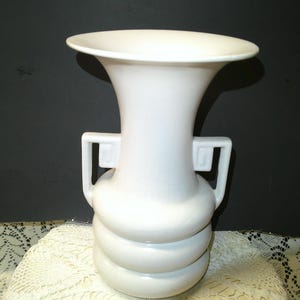 May include: A white ceramic vase with a flared rim and two square handles. The vase has a bulbous base with three stacked, rounded sections. The vase is a solid, matte white color and stands on a lace-covered surface.