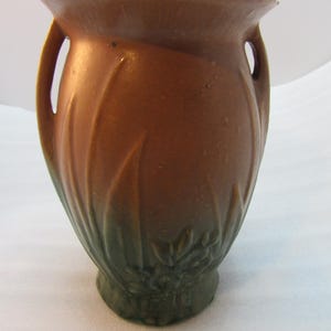 May include: A brown ceramic vase with two handles and a flared rim. The vase has a textured surface with a leaf-like pattern and a dark green base. The vase is approximately 8 inches tall.