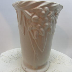 May include: A light pink ceramic vase with a flared top and a round base. The vase features embossed floral and leaf designs. The date "10/22/2025" is printed on the bottom right corner of the image. The vase is sitting on a white lace tablecloth.