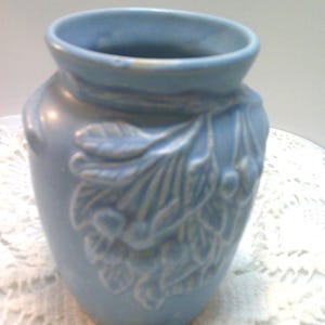 May include: A light blue ceramic vase with a wide opening and a rounded body. The vase features a raised floral design on the front. The vase is sitting on a white lace surface. The vase is approximately 5 inches tall.