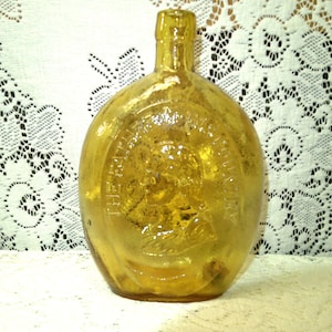 May include: A vintage, amber-colored glass bottle with a rounded body and a long neck. The bottle has an embossed design on the front, likely a logo or emblem. The bottle is displayed on a white lace tablecloth.