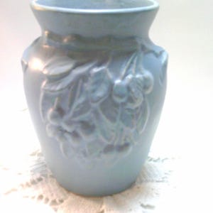 May include: A light blue ceramic vase with a flared top and a detailed embossed cherry blossom design. The vase is approximately 6 inches tall and has a rounded body. The vase is sitting on a white lace doily.