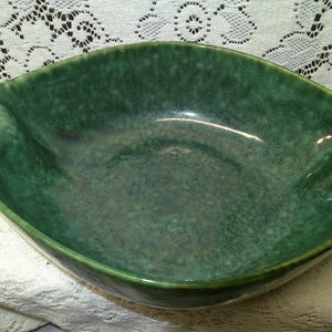 May include: A green ceramic serving bowl with a glossy finish. The bowl has a unique, asymmetrical shape with gently curved sides. The interior has a speckled texture, and the exterior is a solid, deep green color. This decorative bowl is suitable for serving food.