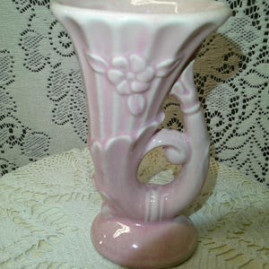 May include: A vintage pink ceramic vase with a flared top and a rounded base. The vase features a floral design and a curved handle. The vase is approximately 8 inches tall and is set against a lace background.
