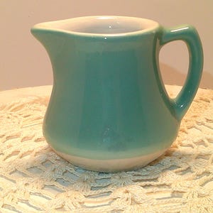 May include: A small, turquoise ceramic pitcher with a white interior and a curved handle. The pitcher has a classic design with a spout for easy pouring. The base is white, and the exterior is a solid turquoise color.