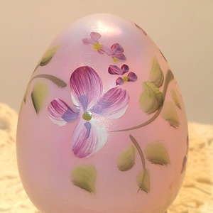May include: A light pink, egg-shaped decorative item with hand-painted floral designs. The flowers are primarily purple and white, with green leaves and stems. The egg has a smooth, glossy finish, and the overall aesthetic is delicate and artistic.