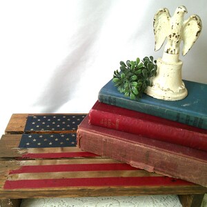 May include: A rustic wooden step stool with a distressed American flag design. A stack of vintage books in red and blue tones supports a cream-colored eagle statue and a small green plant. The overall aesthetic is vintage and patriotic.