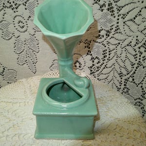 May include: A vintage-style, mint green ceramic vase. The vase is shaped like an old gramophone, with a square base, a round opening, and a flared horn. The vase is a solid color and has a glossy finish. The background is a white lace tablecloth.