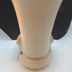 May include: A cream-colored ceramic vase with a flared top and a rounded base. The vase has two decorative elements on the sides and a textured, ridged base. The date "12/03/2025" is printed on the bottom right.