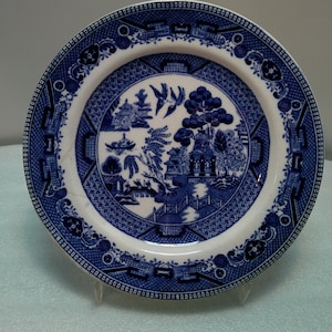 May include: A round, white and blue ceramic plate with a Willow pattern design. The plate features a central scene of a bridge, buildings, and trees, surrounded by a detailed border. The plate is displayed on a clear stand.