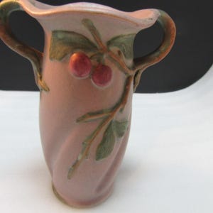 May include: A pink ceramic vase with two handles and a decorative cherry branch design. The vase has a twisted body and is adorned with red cherries and green leaves. The handles are brown and green.
