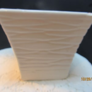 May include: A small, off-white square ceramic planter with a textured, horizontal wave pattern. The planter has a trapezoidal shape, wider at the top and narrower at the base. The planter is approximately 4 inches tall.