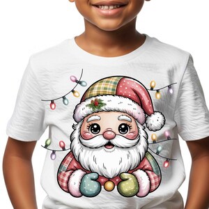 Peut inclure: T-shirt blanc avec un dessin de Père Noël de dessin animé. Le Père Noël a une barbe blanche, des joues roses et un chapeau rouge et à carreaux. Des guirlandes lumineuses colorées entourent l'illustration du Père Noël. Le t-shirt est porté par un enfant.