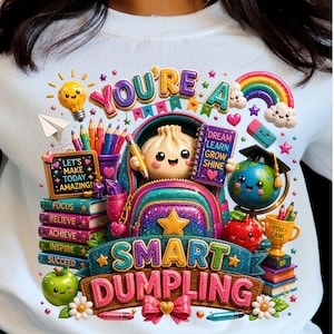 Peut inclure: Un sweat-shirt blanc avec un motif coloré représentant une boulette souriante dans un sac à dos, entourée de fournitures scolaires, d'un globe, d'un arc-en-ciel et de livres. Le texte indique : YOU'RE A SMART DUMPLING, LET'S MAKE TODAY AMAZING, FOCUS, BELIEVE, ACHIEVE, INSPIRE, SUCCEED, DREAM LEARN GROW SHINE, et YOU CAN!