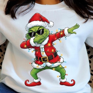 Puede incluir: Sudadera blanca con un personaje de dibujos animados verde con un traje de Papá Noel y gafas de sol, haciendo un dab. El personaje está envuelto en luces navideñas y lleva un gorro y zapatos rojos de Papá Noel. El diseño es festivo y humorístico.