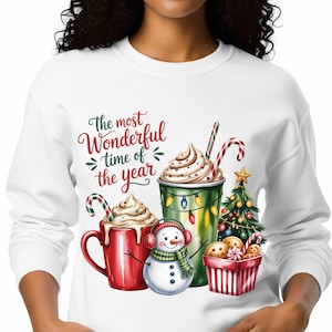 Peut inclure: Sweat-shirt blanc avec un motif festif. Le motif présente une tasse rouge et un gobelet vert de boissons à la crème fouettée, un bonhomme de neige, un sapin de Noël et un panier de biscuits. Le texte dit : "The most Wonderful time of the year."