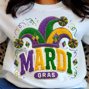 Peut inclure: Sweat-shirt blanc avec un motif Mardi Gras. Le motif représente un chapeau de bouffon violet, vert et jaune, avec le mot "MARDI" en grandes lettres et "GRAS" en dessous. Le motif comprend des paillettes dorées et des volutes décoratives.