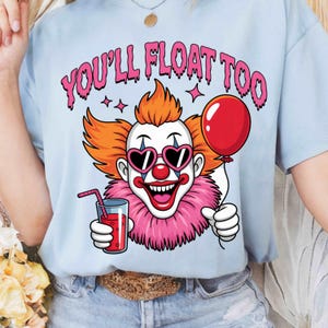 Könnte beinhalten: Hellblaues T-Shirt mit einem Cartoon-Clown-Design. Der Clown hat orangefarbene Haare, herzförmige Sonnenbrillen und einen rosa Rüschenkragen. Er hält einen roten Ballon und ein Glas rote Flüssigkeit mit einem Strohhalm. Der Text "YOU'LL FLOAT TOO" steht über dem Clown.