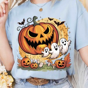 Peut inclure: T-shirt bleu clair avec un motif sur le thème d'Halloween. Le dessin comprend une grande citrouille orange avec un visage sculpté, trois fantômes blancs, des chauves-souris et de plus petites citrouilles. Le fond présente des étoiles et un cercle jaune.