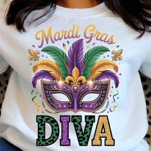 May include: White sweatshirt with a Mardi Gras design. The design features a purple and gold mask with green, purple, and gold feathers. The words "Mardi Gras" and "DIVA" are also printed on the sweatshirt.