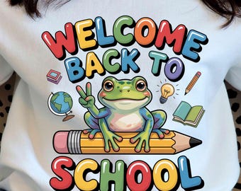 May include: White sweatshirt with a colorful graphic that says "WELCOME BACK TO SCHOOL". The design features a friendly frog on a pencil, with school-themed illustrations. The text is in a rainbow of colors.