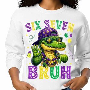 Peut inclure: Sweat-shirt blanc avec un motif d'alligator vert portant des lunettes de soleil, une casquette violette et un collier de perles. Le texte "SIX SEVEN BRUH" est imprimé au-dessus et en dessous de l'alligator.