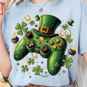 Peut inclure: T-shirt bleu clair avec un motif de manette de jeu verte ornée d'un chapeau de leprechaun, de trèfles et de pièces d'or. La manette comporte des boutons avec des lettres et un pavé directionnel. Un arc-en-ciel et des fers à cheval complètent le design de la Saint-Patrick.