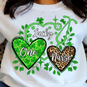 Peut inclure: Sweat-shirt blanc avec un motif de la Saint-Patrick. Le graphisme comprend un cœur vert avec des trèfles et le mot "One", un cœur léopard avec le mot "Nurse", et le mot "Lucky" en vert. Un stéthoscope et des trèfles complètent le design.